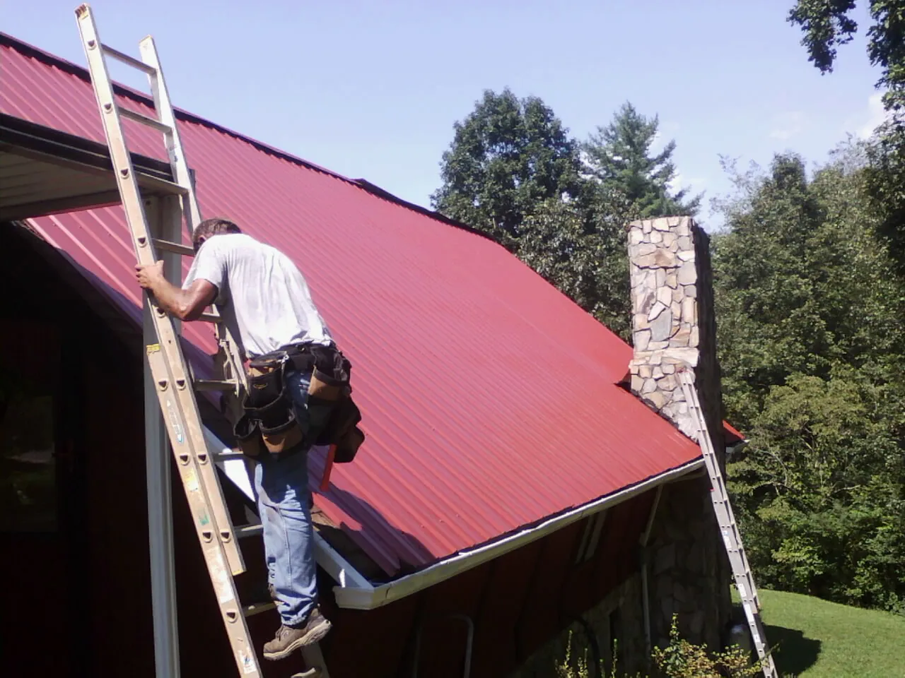 Roofer on a ladder beside a red metal roof and stone chimney — Soffit & Fascia Repair by Roofing Brothers in Ashville